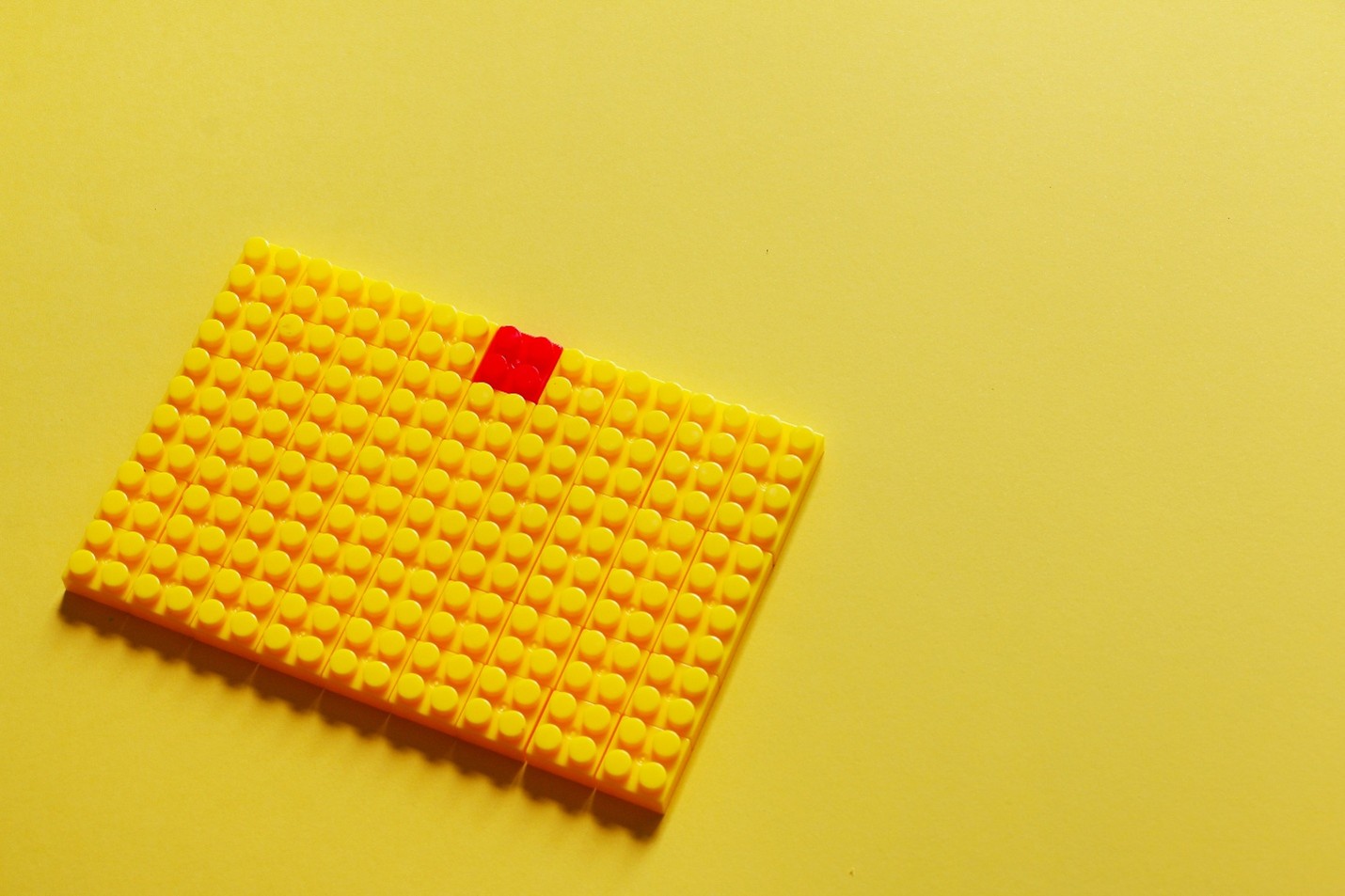 A rectangle of yellow Lego 2x2 bricks with a red Lego 2x2 brick in the center of the top row on a background of yellow