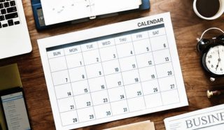 A calendar, clock, computer, phone, and tablet on a wooden table to help complete reports on time